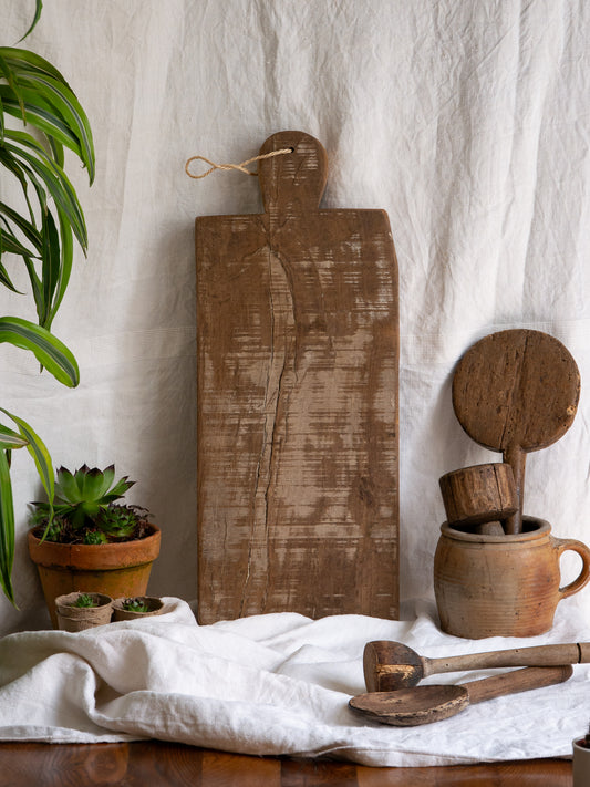 A large vintage wooden chopping board from south west France, shaped by years of use and hand-worn edges. Its rough texture and deep patina tell the story of daily bread, shared meals, and time well lived.