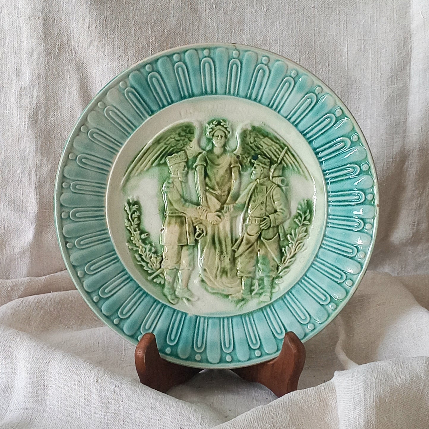 A French antique decorative majolica plate with stunning blue/green colours and an interesting historical background.
This lovely old plate was manufactured as a commemorative piece to celebrate the Franco-Russian Alliance made by the French Republic and the Russian Empire between 1891-1894 (military co-operation agreements) – and that lasted until 1917. The plate depicts a Russian and French soldier shaking hands with a winged female angel in between them.