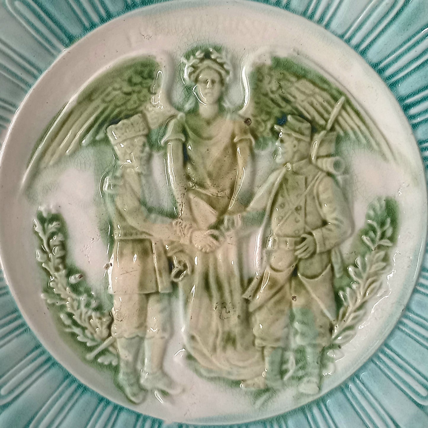 A French antique decorative majolica plate with stunning blue/green colours and an interesting historical background.
This lovely old plate was manufactured as a commemorative piece to celebrate the Franco-Russian Alliance made by the French Republic and the Russian Empire between 1891-1894 (military co-operation agreements) – and that lasted until 1917. The plate depicts a Russian and French soldier shaking hands with a winged female angel in between them.