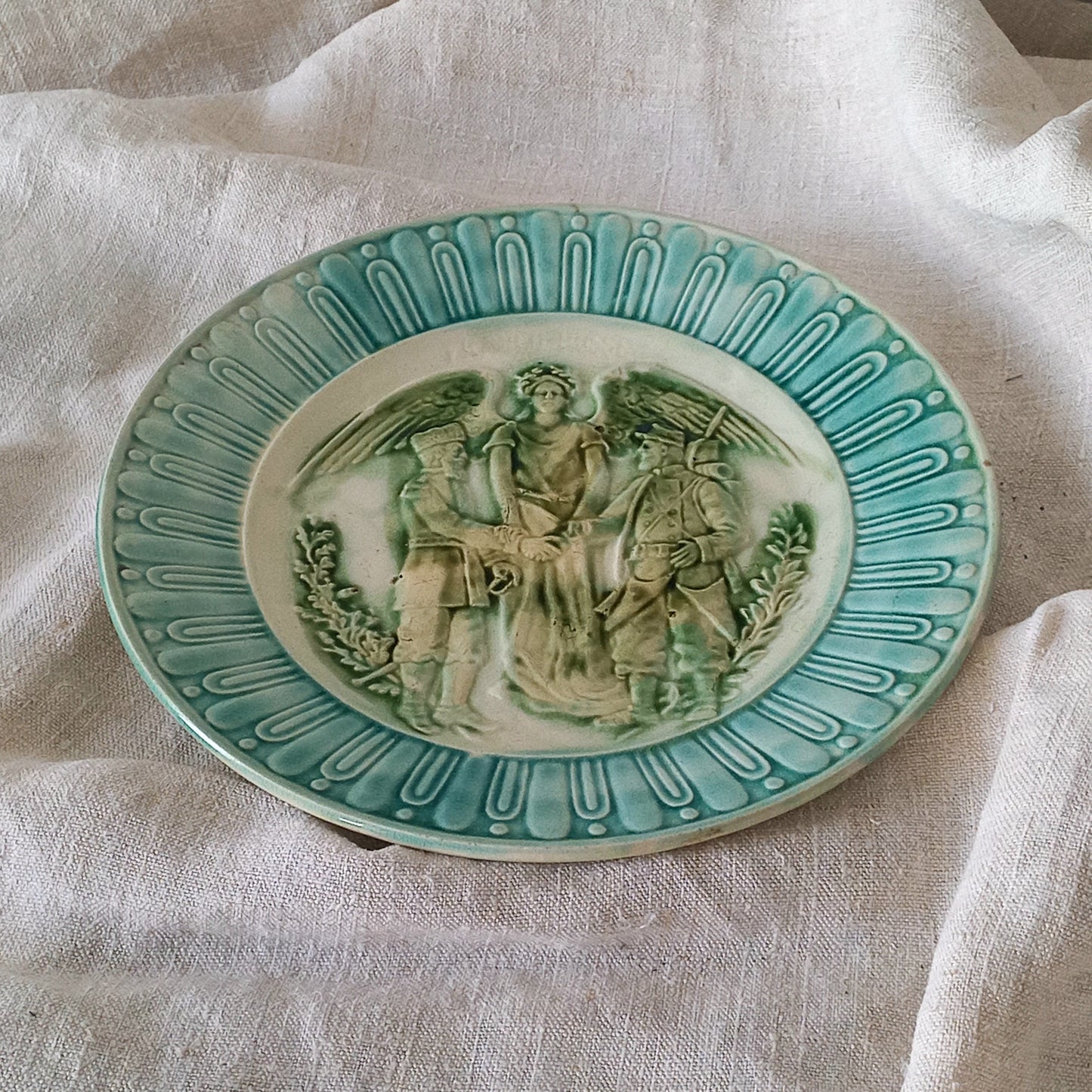 A French antique decorative majolica plate with stunning blue/green colours and an interesting historical background.
This lovely old plate was manufactured as a commemorative piece to celebrate the Franco-Russian Alliance made by the French Republic and the Russian Empire between 1891-1894 (military co-operation agreements) – and that lasted until 1917. The plate depicts a Russian and French soldier shaking hands with a winged female angel in between them.