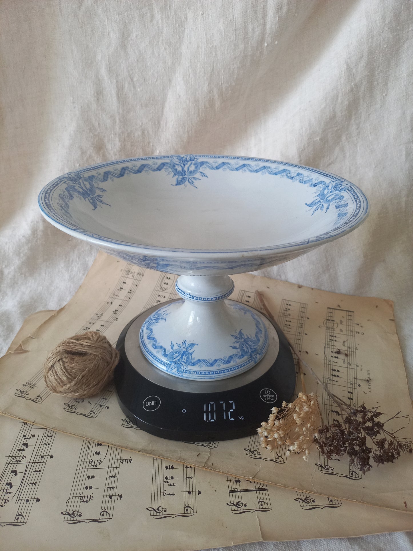 A white ceramic pedestal bowl with blue floral patterns sits on a digital scale placed on vintage sheet music, with twine and dried flowers nearby against a draped fabric backdrop.