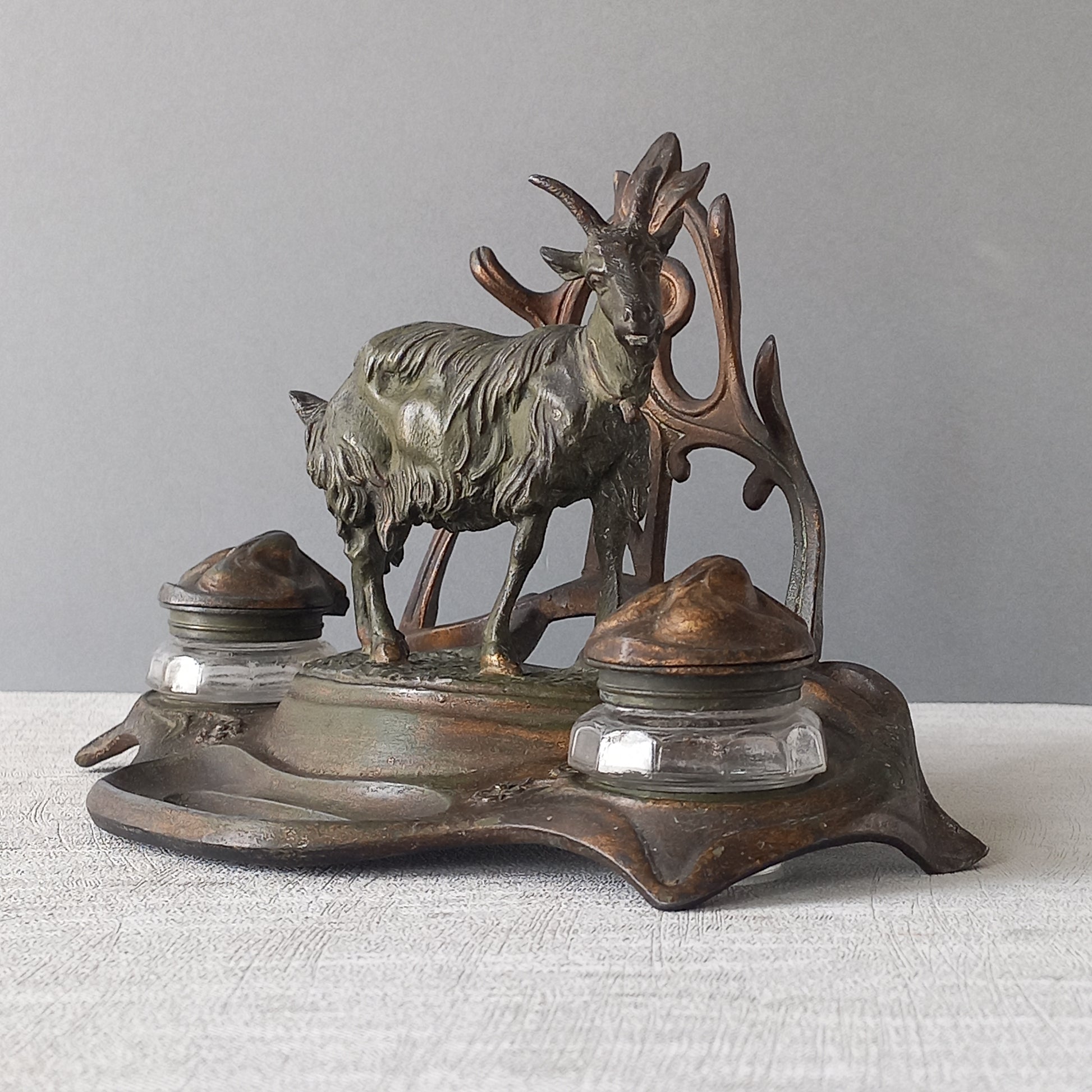 A stunning French art nouveau double inkwell with a goat figurine.
This wonderful antique inkwell has been sculpted in metal and signed by the artist (see photos). Behind the beautiful goat figurine are two pen rests and either side of the goat are two small glass ink pots with decorative metal lids.
The metal is finished in a lovely shade of green that has worn over time with traces of gold colour beneath - it has a wonderful patina of time all over it.