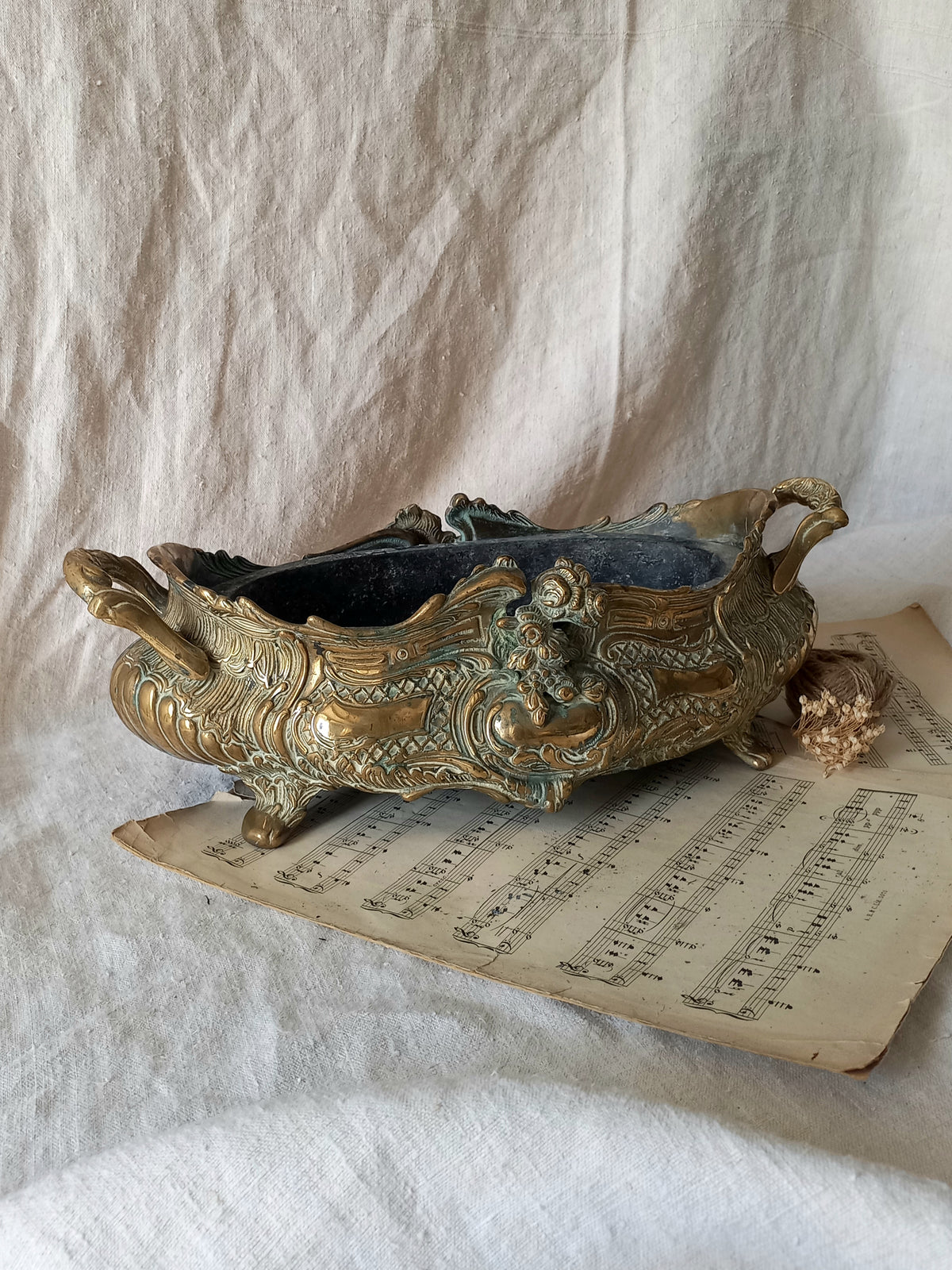 An ornate antique decorative planter resting on a vintage sheet music page against a draped white fabric background.