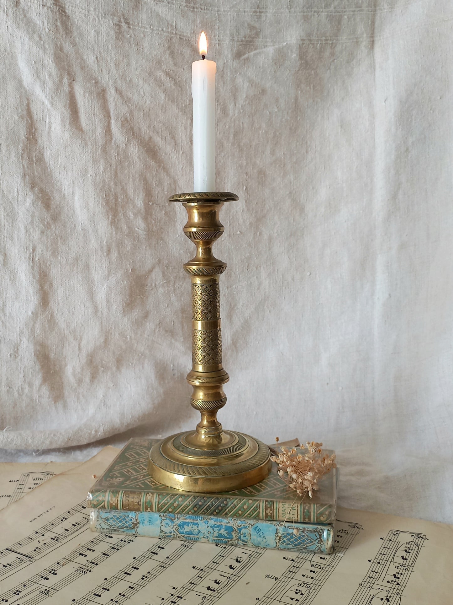 An ornate brass candlestick holder with a lit white taper candle, displayed on top of vintage books over sheet music against a draped fabric backdrop.