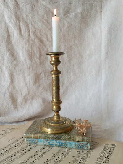 An ornate brass candlestick holder with a lit white taper candle, displayed on top of vintage books over sheet music against a draped fabric backdrop.
