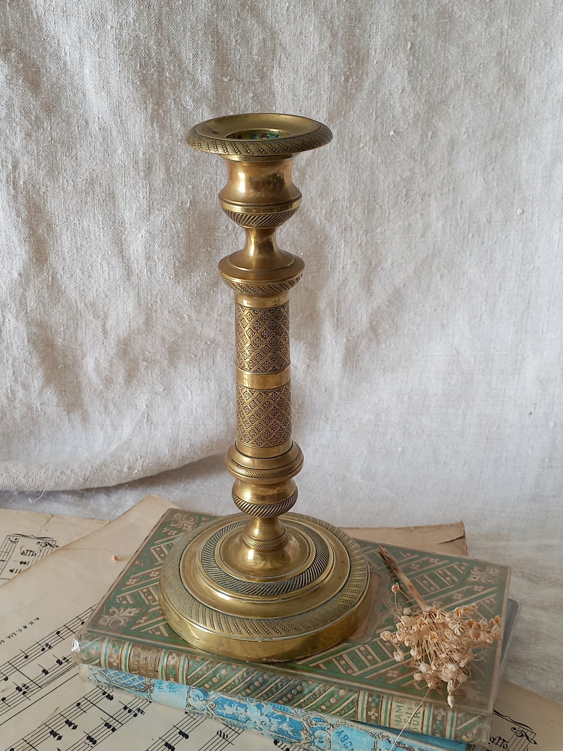 An ornate vintage brass candlestick holder with intricate textured detailing, displayed on top of a decorative book with sheet music underneath against a draped white fabric background.