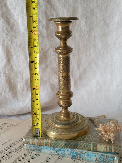 An antique brass candlestick holder with a textured grip, displayed next to a yellow tape measure to show its height, resting on a vintage book with a small dried flower on a white fabric background.