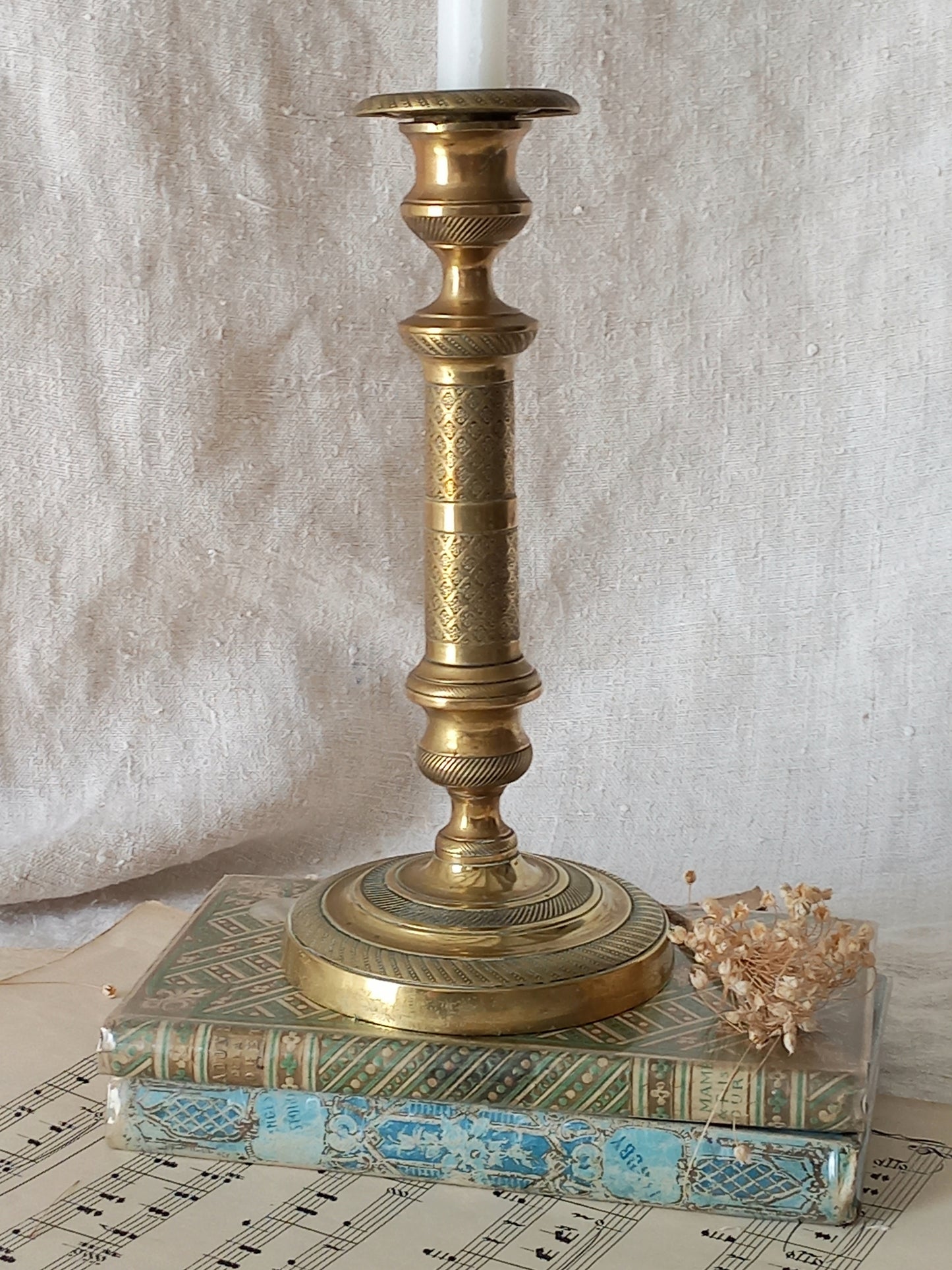An ornate vintage brass candlestick holder with a carved column and round base, holding a white candle, styled on top of two antique books with a small dried floral sprig against a draped fabric backdrop.