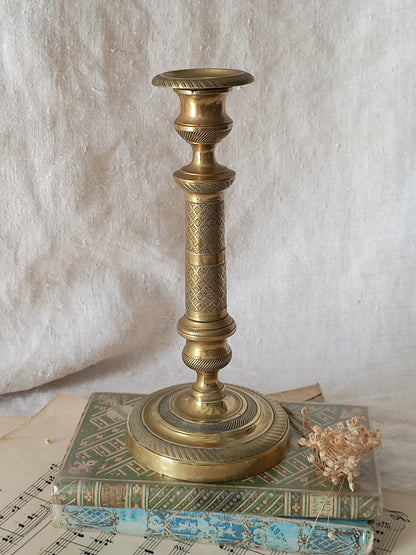 An ornate vintage brass candlestick holder with intricate geometric engravings, displayed on top of two antique books against a textured off-white fabric backdrop.