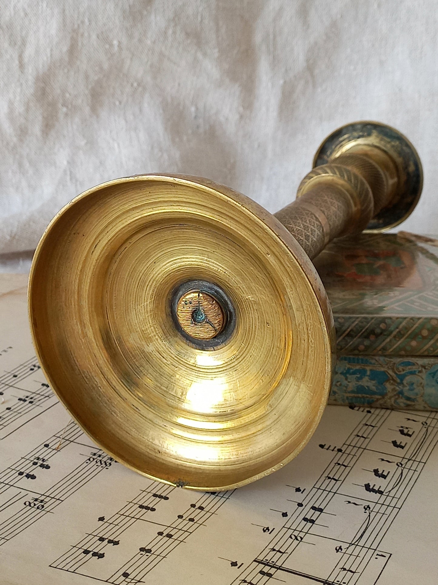 A close-up of the underneath of a brass candle holder, resting on printed sheet music with a textured white fabric background.