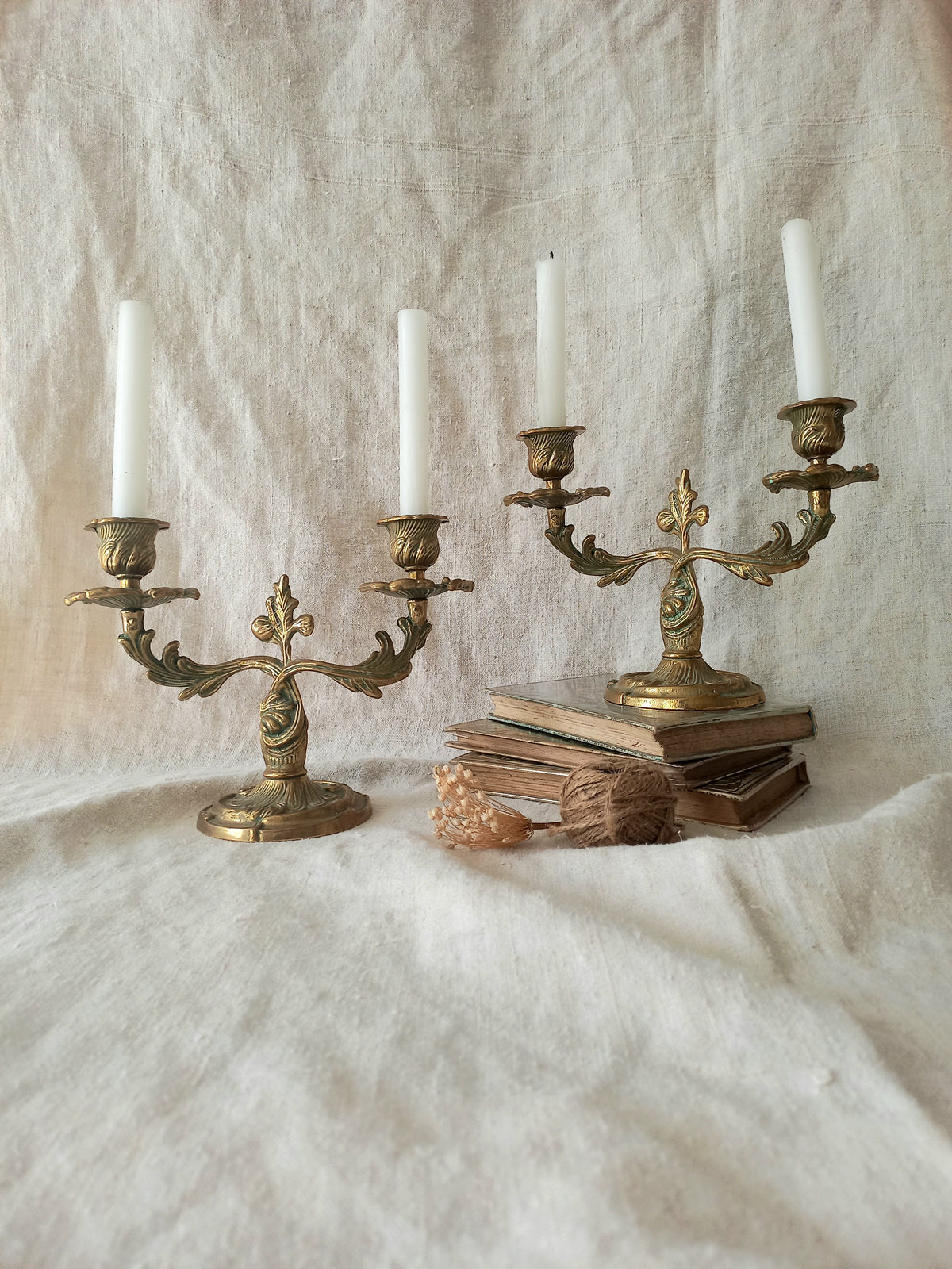 Two ornate brass candelabras with white candles, styled on a draped cream linen fabric with vintage books and a small decorative object.