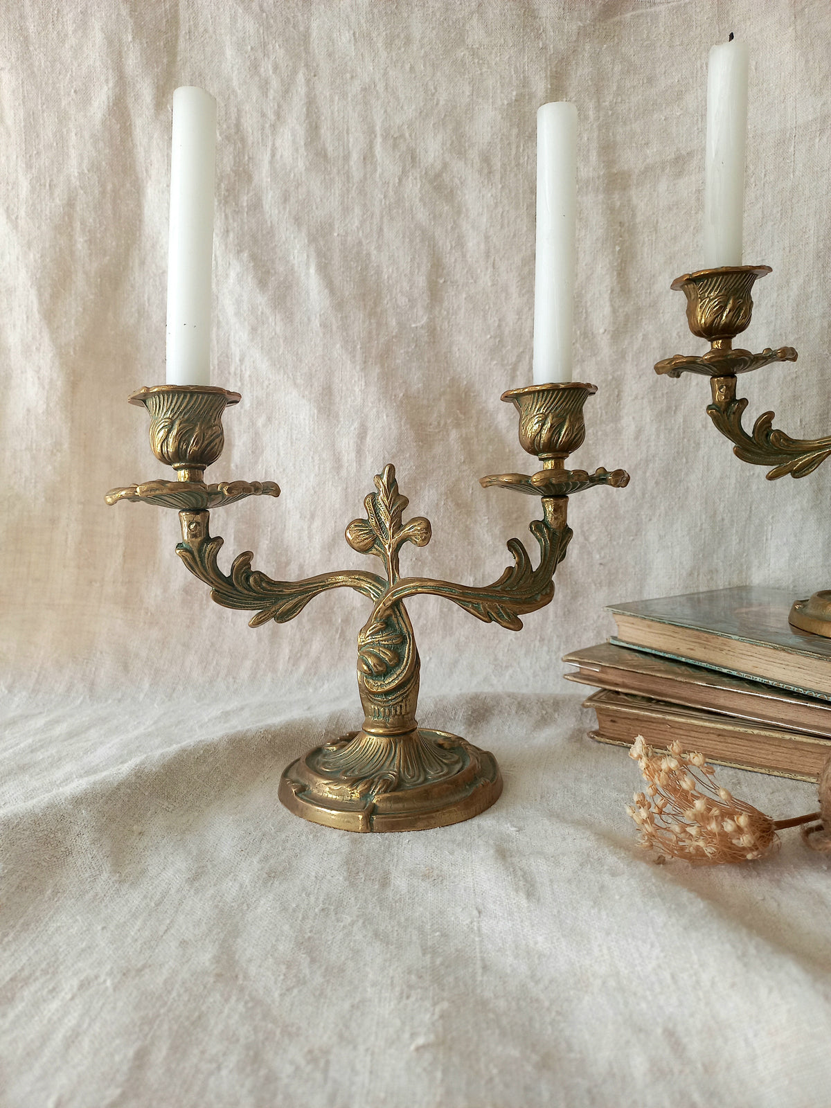 An ornate brass candelabra with two white taper candles, styled on a draped beige fabric surface next to vintage books and a small dried floral sprig.