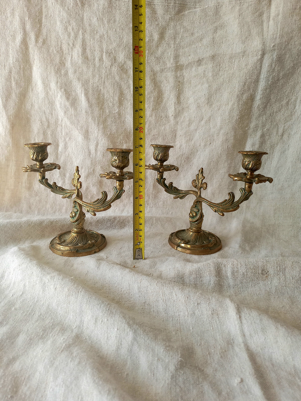 A pair of ornate brass candelabras displayed side-by-side on a wrinkled white fabric backdrop, with a yellow tape measure standing vertically between them for scale.