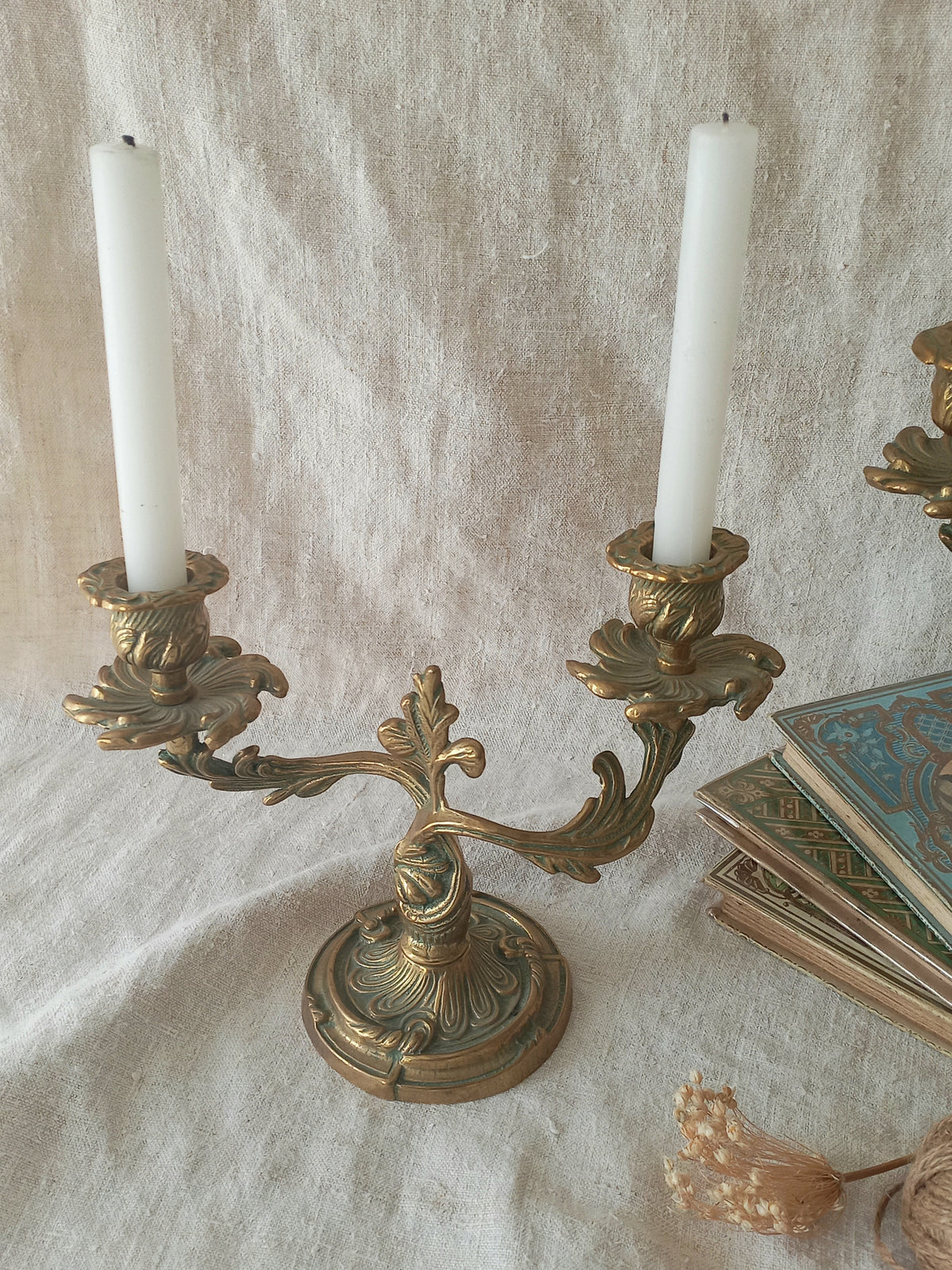 An ornate antique-style brass candelabra with two white taper candles, set against a draped beige linen fabric backdrop with vintage books and dried flowers as props.