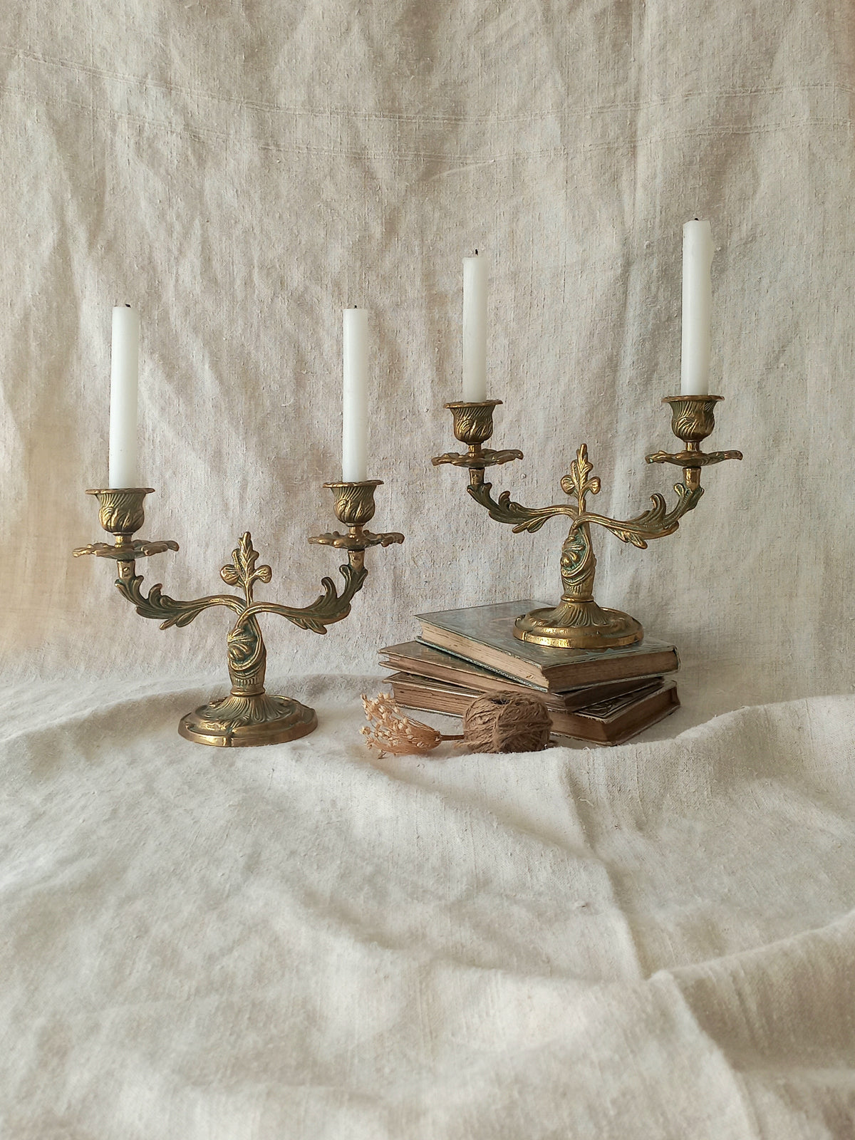 Two ornate antique brass candelabras with white taper candles, styled on a draped cream linen cloth with vintage books and a small shell.