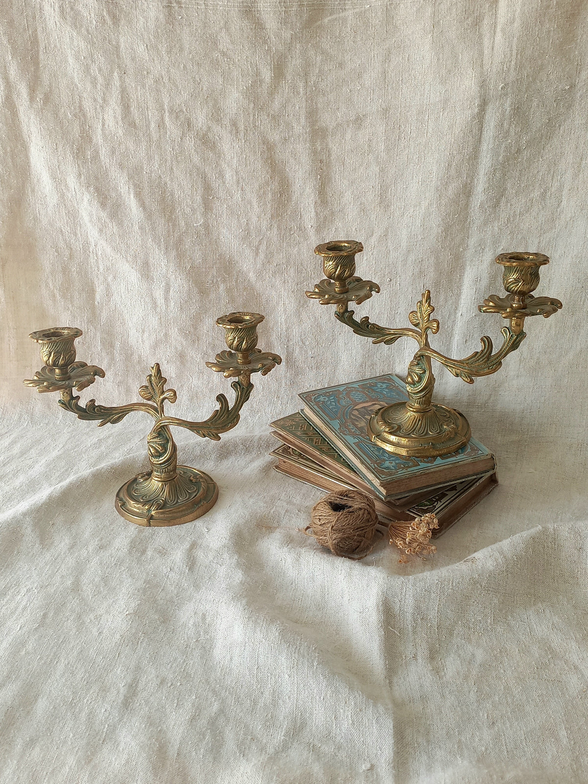 Two ornate brass candelabras displayed on draped white linen fabric, with one holding a stack of vintage books and a small dried flower.