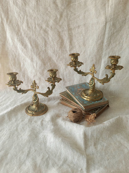 Two ornate brass candelabras displayed on draped white linen fabric, with one holding a stack of vintage books and a small dried flower.