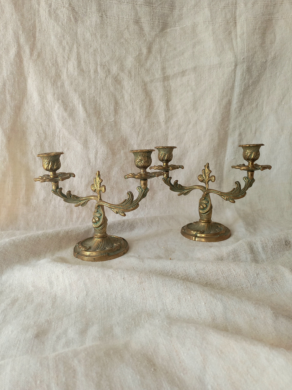 A pair of ornate brass candelabras with three arms each, displayed on a draped cream linen fabric background.