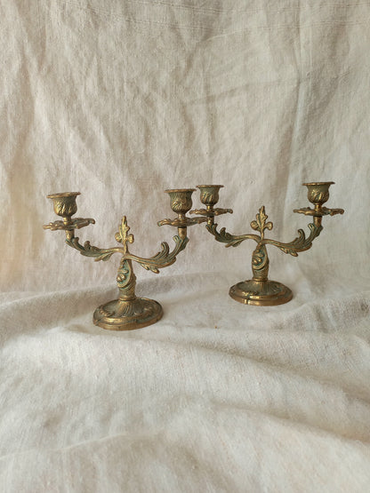 A pair of ornate brass candelabras with three arms each, displayed on a draped cream linen fabric background.