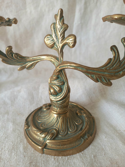 An ornate brass candelabra with a circular pedestal base and leaf-shaped arms, photographed against a draped white fabric background.