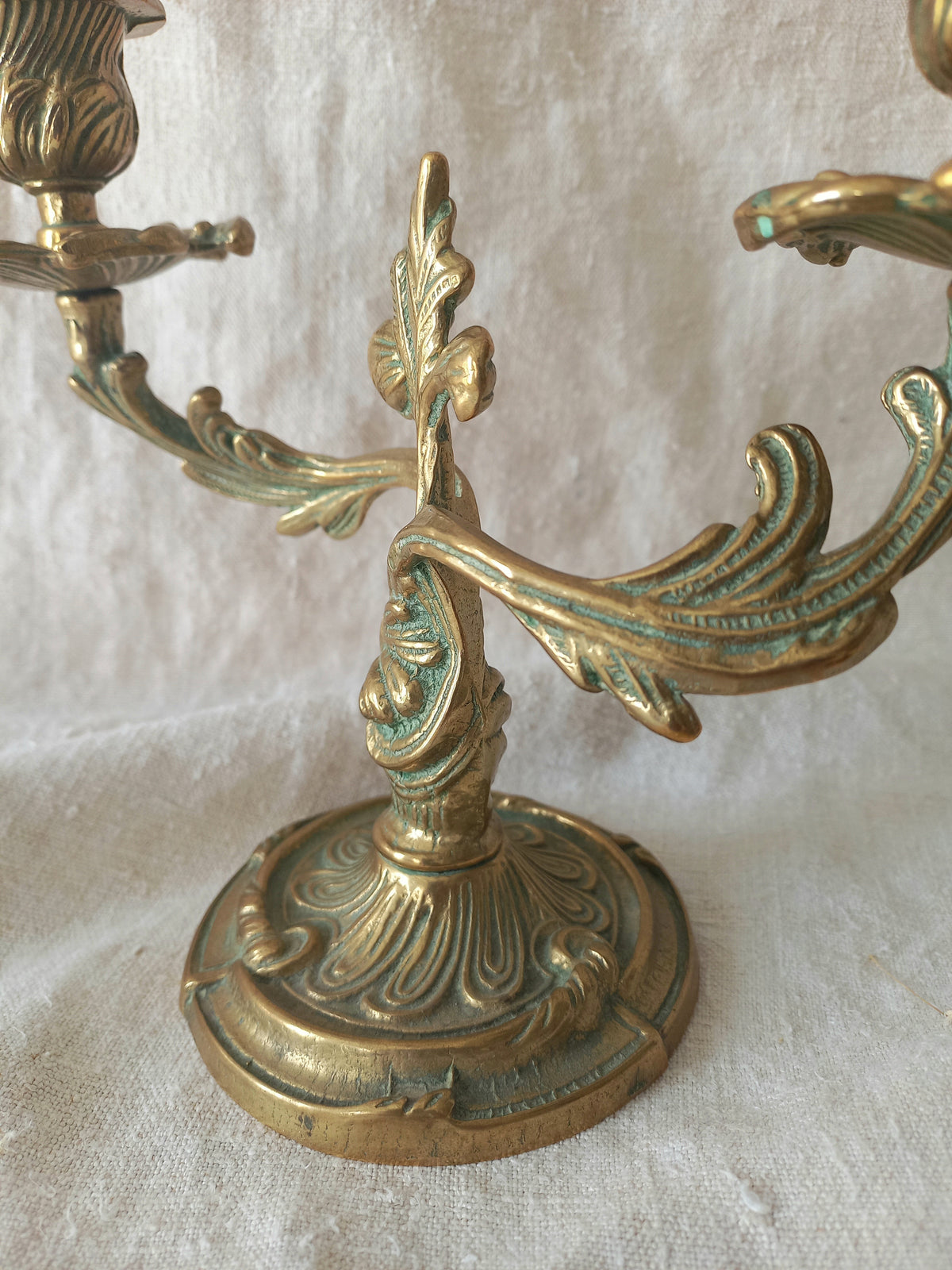 An ornate brass candelabra with a distressed greenish patina, featuring leafy scrollwork and a round base, photographed against a textured off-white fabric background.