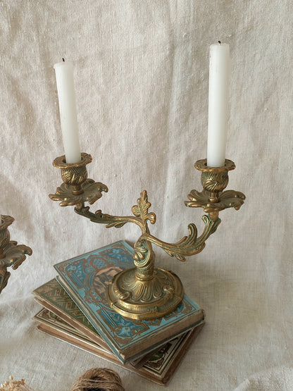 An ornate brass candelabra holding two white candles, styled on top of antique patterned books against a draped linen fabric backdrop.