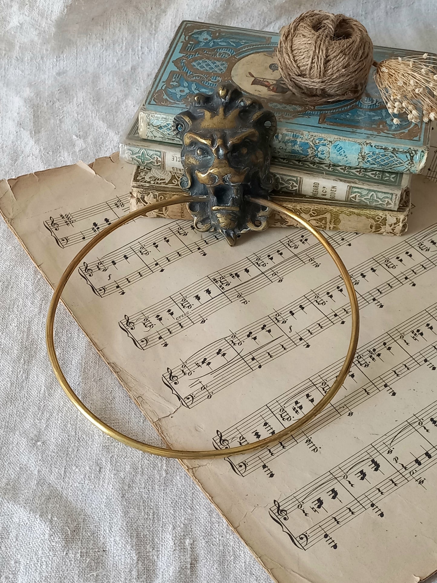 A vintage brass towel ring holder with a lion head sculpted on the front, displayed on top of an antique sheet music page with decorative books and a ball of twine in the background.