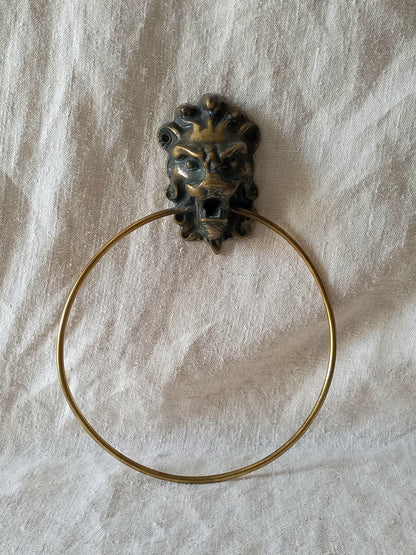 A brass lion head ring holder with a circular ring, displayed on a textured off-white fabric background.