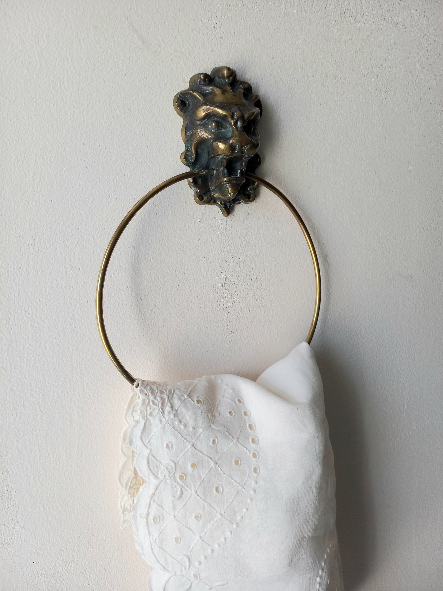 A brass towel ring with a decorative lion head finial, holding a white embroidered cotton towel against a plain off-white wall.