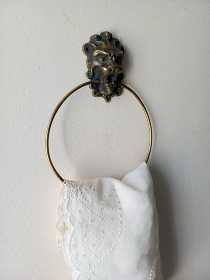 A brass towel ring with a decorative lion head finial, holding a white embroidered cotton towel against a plain off-white wall.