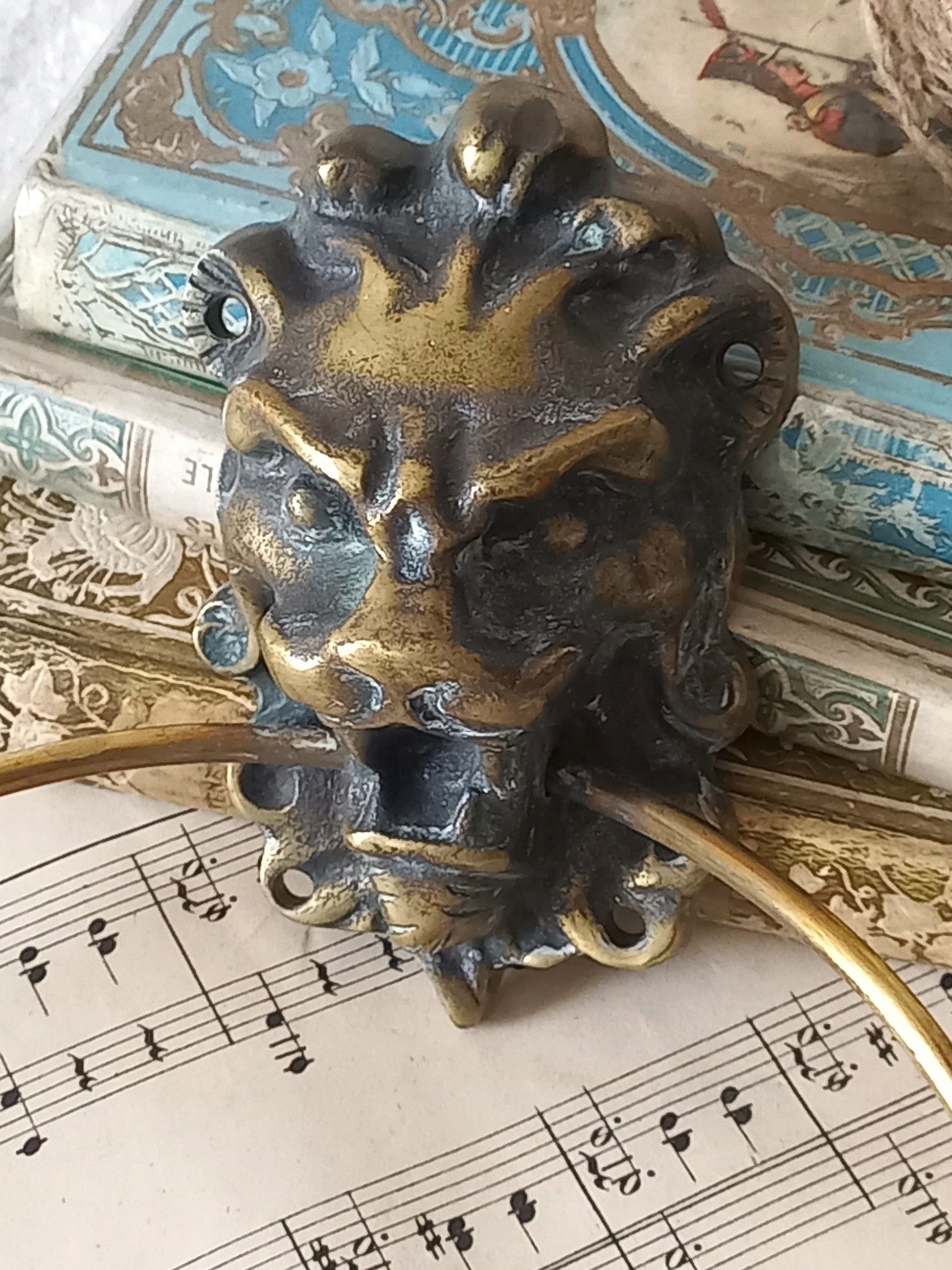 A vintage brass towel ring holder with a lion head sculpted on the front, displayed on top of an antique sheet music page with decorative books in the background.