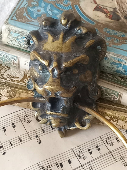 A vintage brass towel ring holder with a lion head sculpted on the front, displayed on top of an antique sheet music page with decorative books in the background.