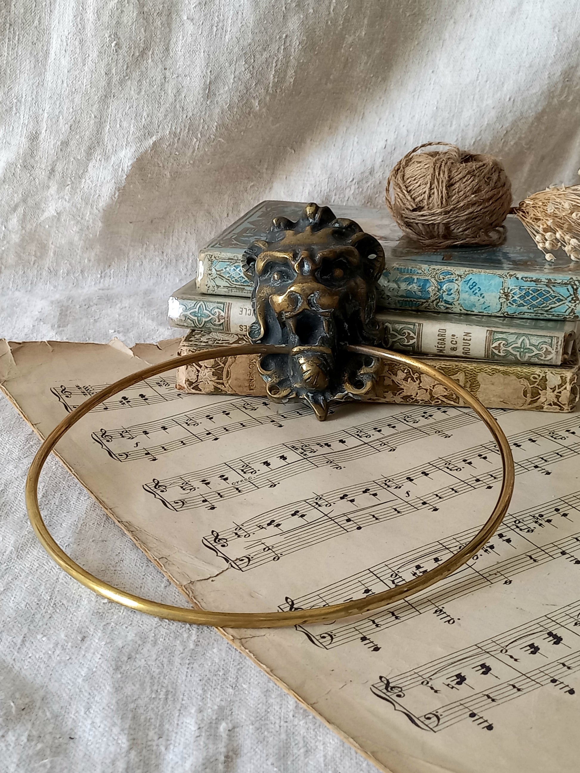 A vintage brass towel ring holder with a lion head sculpted on the front, displayed on top of an antique sheet music page with decorative books and a ball of twine in the background.
