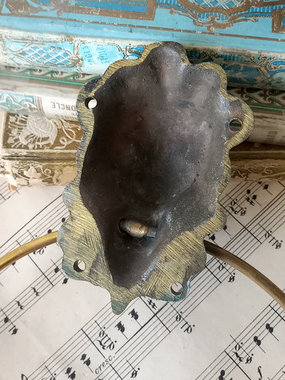 Close-up of rear of a vintage brass towel ring holder with a lion head sculpted on the front, displayed on top of an antique sheet music page with decorative books in the background.