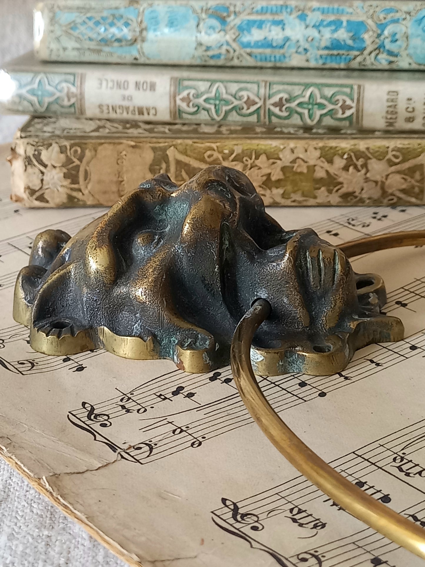Close-up of rear of a vintage brass towel ring holder with a lion head sculpted on the front, displayed on top of an antique sheet music page with decorative books  in the background.