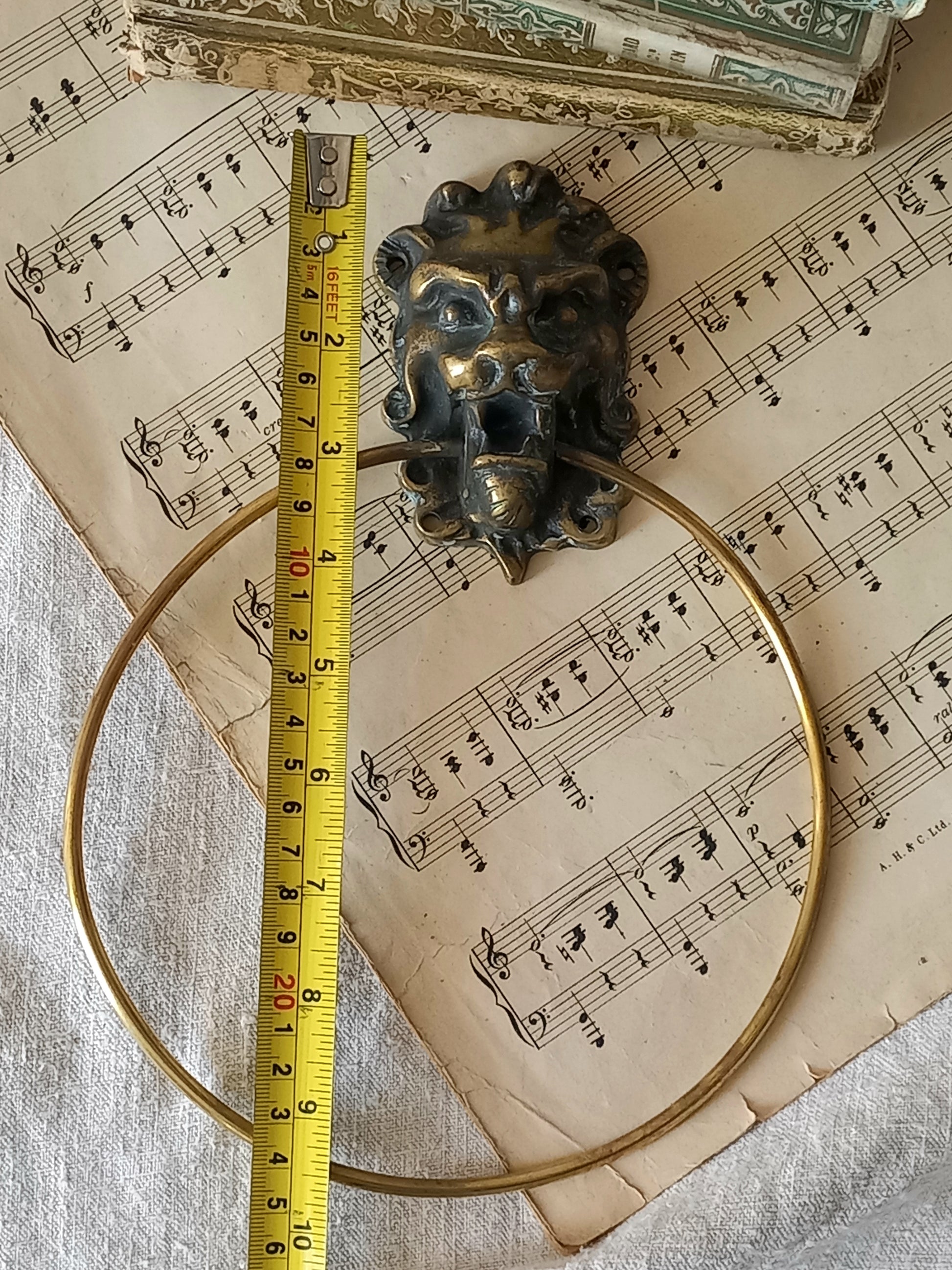 vintage brass towel ring holder with a lion head sculpted on the front, displayed on top of an antique sheet music page with decorative books in the background with tape measure for reference.