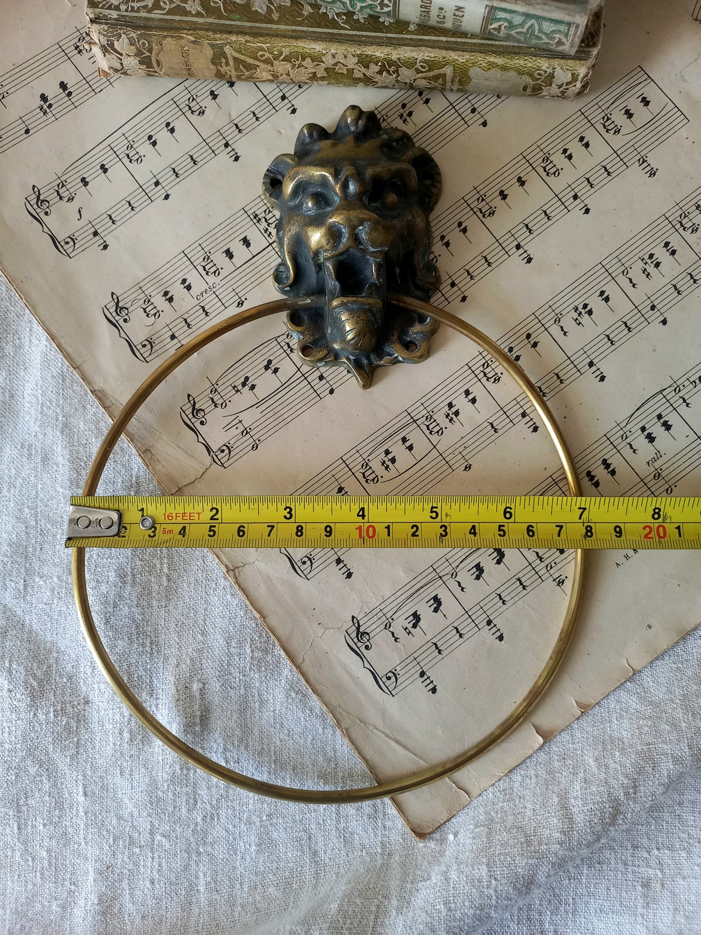 vintage brass towel ring holder with a lion head sculpted on the front, displayed on top of an antique sheet music page with decorative books in the background with tape measure for reference.