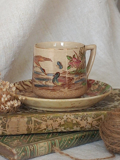 A little antique cup and saucer with oriental influenced design depicting ducks, butterflies, flowers and bamboo shoots, placed on an antique books display.