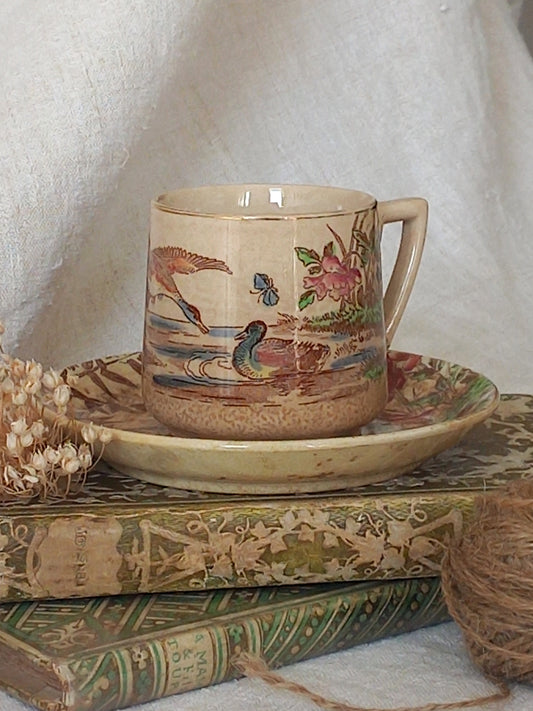 A little antique cup and saucer with oriental influenced design depicting ducks, butterflies, flowers and bamboo shoots, placed on an antique books display.