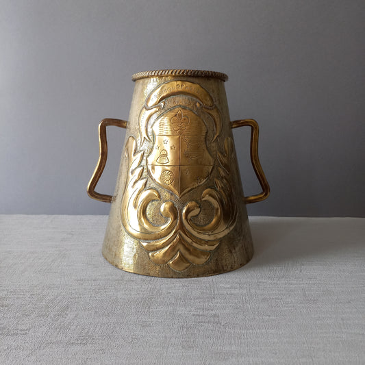 A decorative vintage or antique brass vase or large tankard with two handles. On one side is a coat of arms and on the other an attractive design.