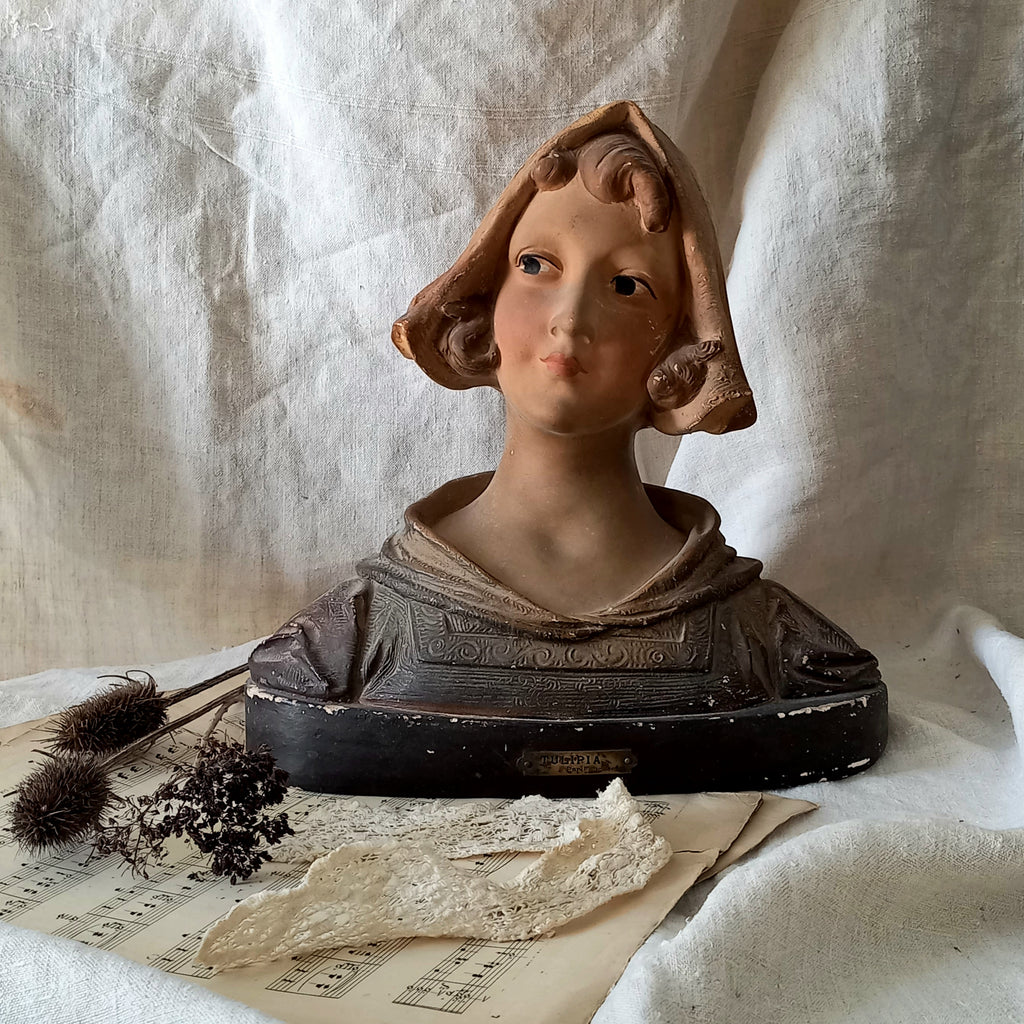Antique bust of a young girl on a textured surface with dried plants and paper.