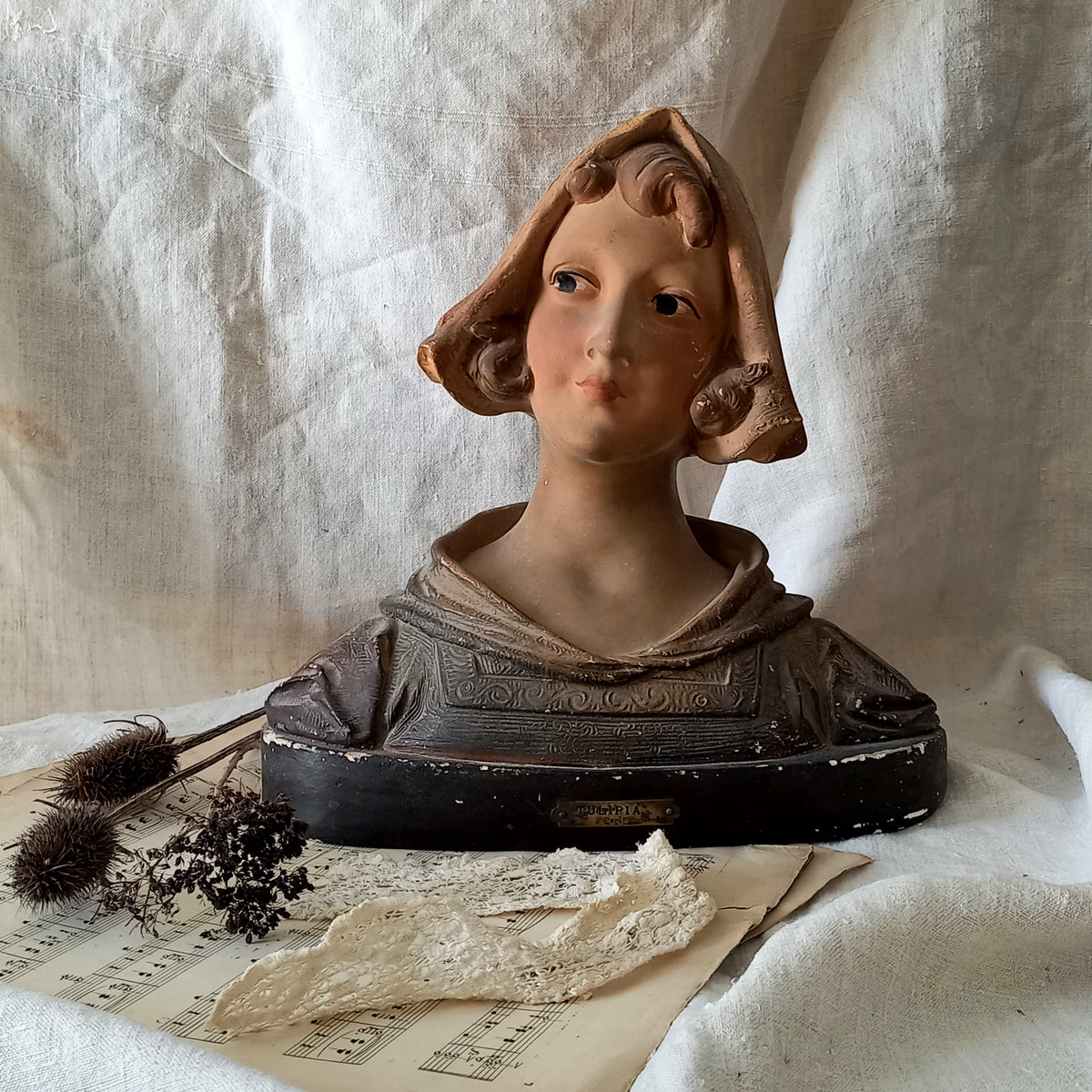 Antique bust of a young girl on a textured surface with dried plants and paper.