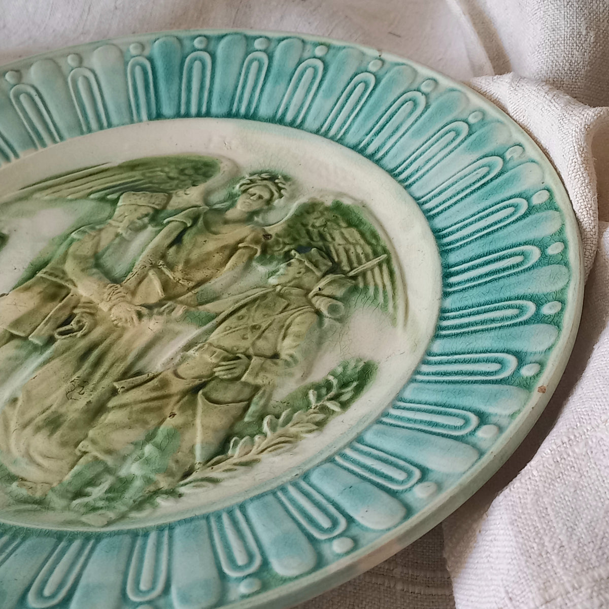 A French antique decorative majolica plate with stunning blue/green colours and an interesting historical background.

This lovely old plate was manufactured as a commemorative piece to celebrate the Franco-Russian Alliance made by the French Republic and the Russian Empire between 1891-1894 (military co-operation agreements) – and that lasted until 1917. The plate depicts a Russian and French soldier shaking hands with a winged female angel in between them.