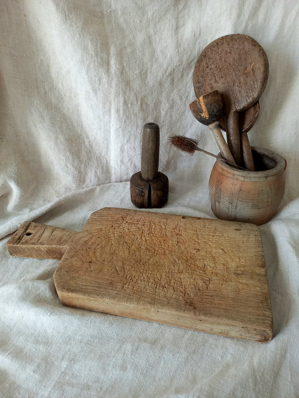 A rustic wooden cutting board with a handle lies on a draped white linen cloth, alongside a dark wooden pestle and a small wooden container holding several wooden spoons.