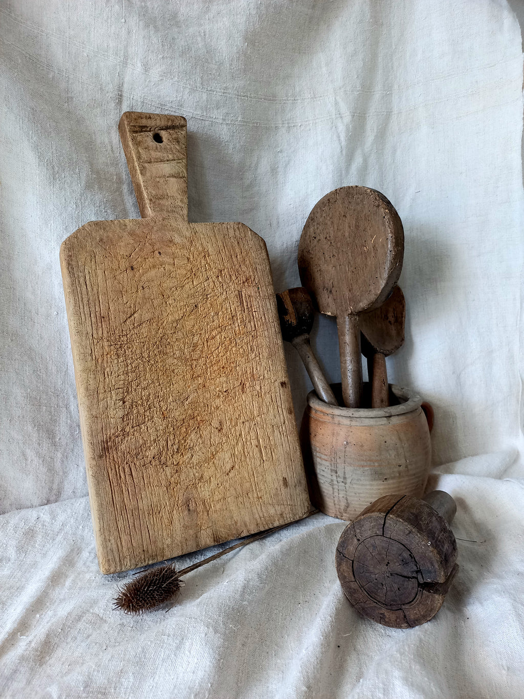 A rustic wooden cutting board with a handle, shown alongside a small ceramic pot holding wooden spoons, a wooden brush, and a wooden mallet on a draped white fabric background.
