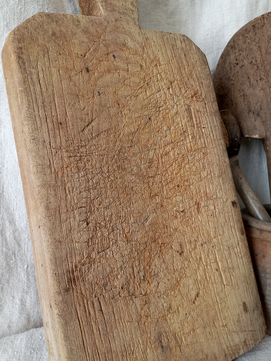 A close-up of a heavily used, rustic wooden cutting board with a handle, showing deep knife scratches and wear, resting on a white textured fabric next to another wooden kitchen tool.