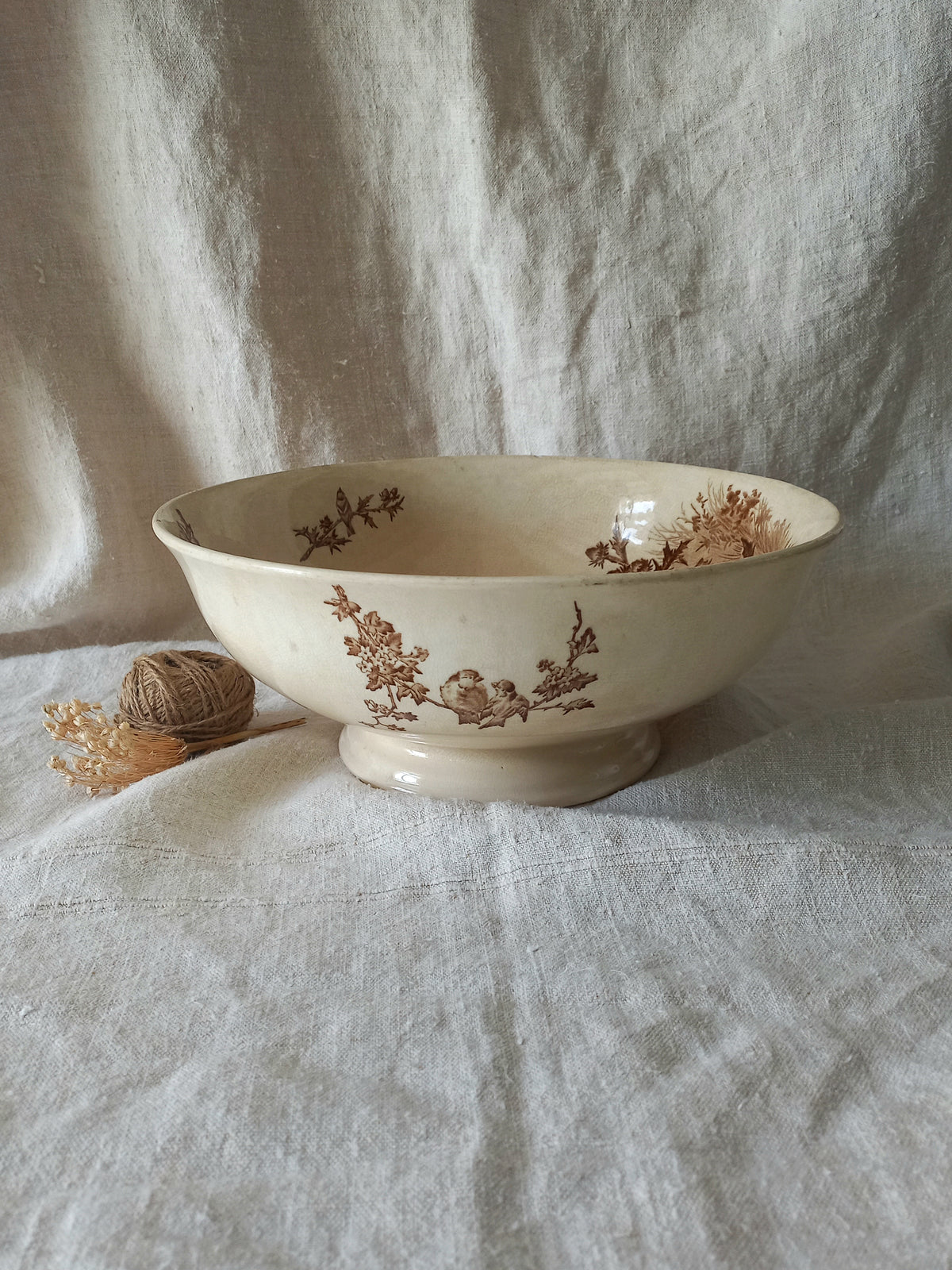 An off-white ceramic bowl with a brown floral toile pattern, set on a draped linen fabric background next to a small decorative ball of twine.