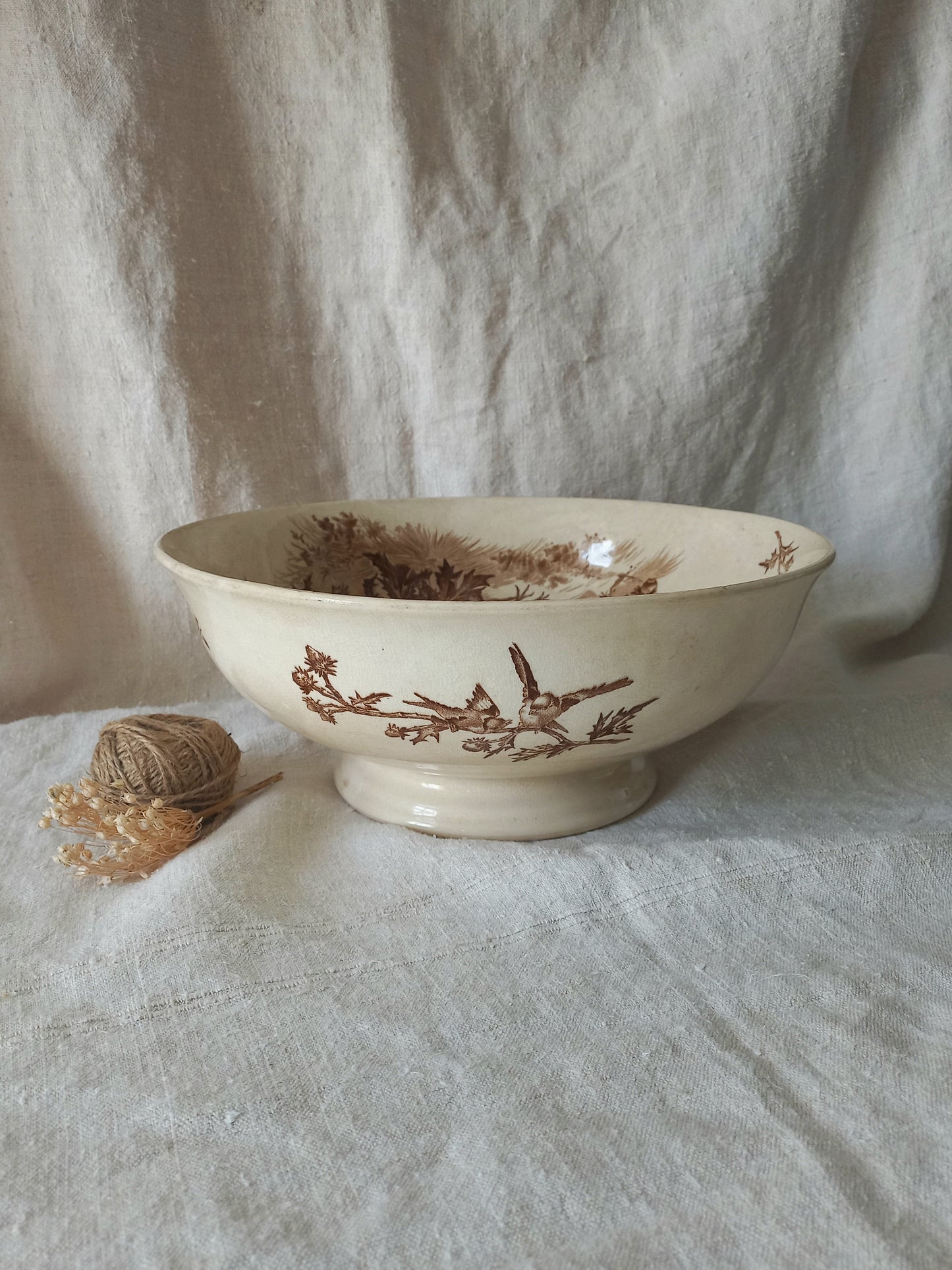 An off-white ceramic bowl with a brown transfer-style print of birds and foliage, set against a draped linen backdrop with a small decorative ball of twine on the left.