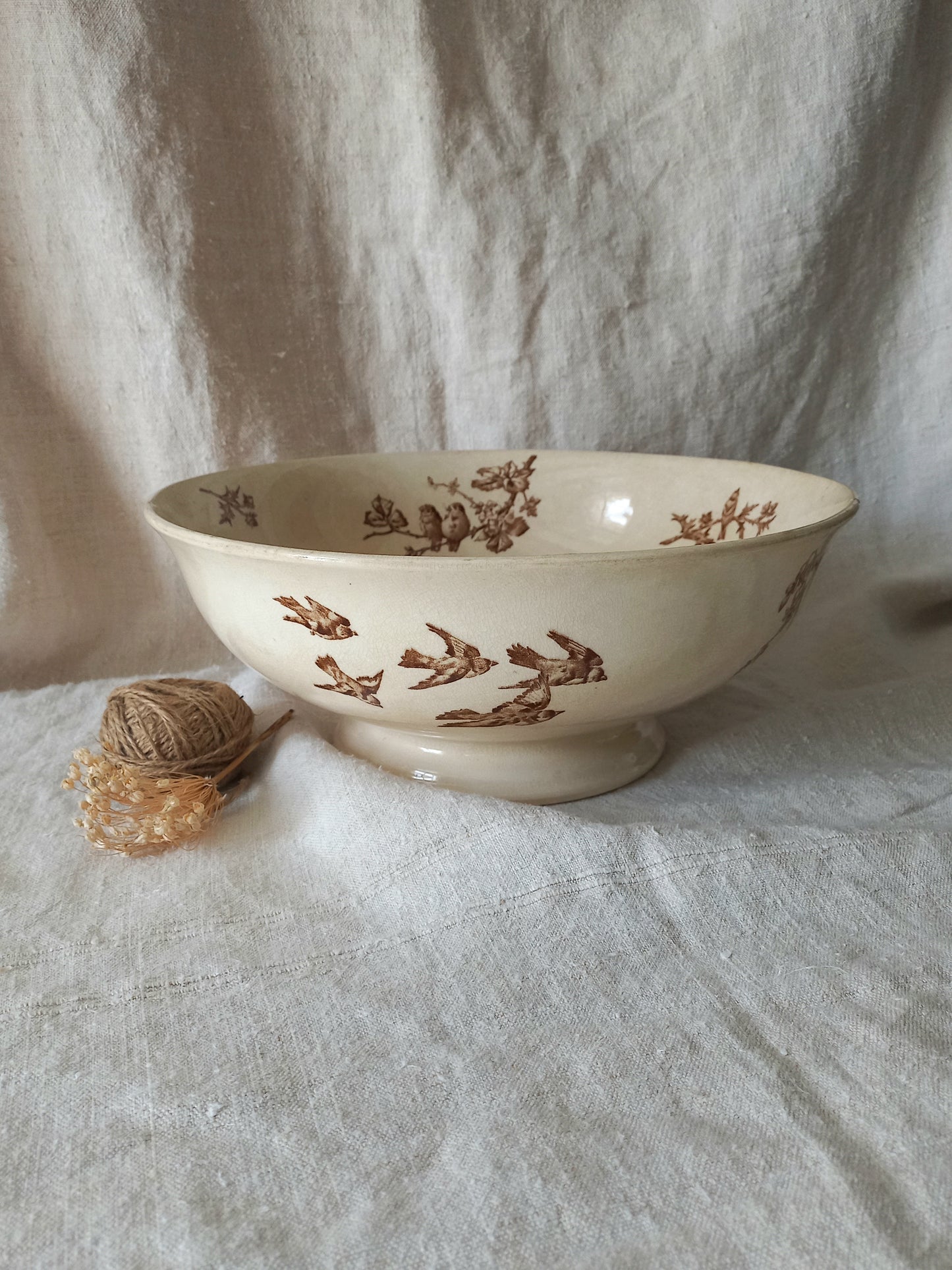 An off-white ceramic bowl with a brown bird and branch pattern, set on a draped linen fabric background next to a small decorative bundle of twine and dried flowers.