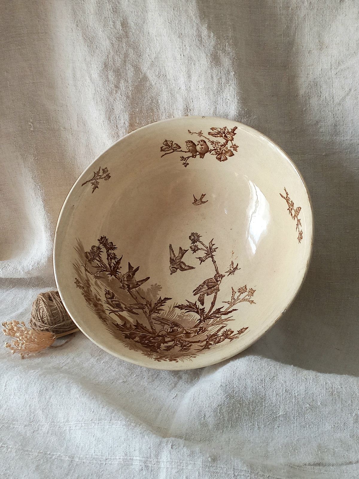 A cream-colored ceramic bowl with a brown toile-style print of birds and foliage, displayed on draped white linen fabric.
